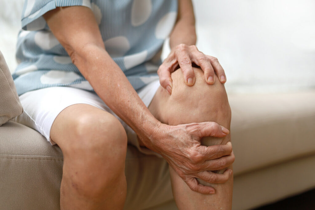 A senior Asian woman is massaging her knee while sitting on the sofa, suffering from knee pain.