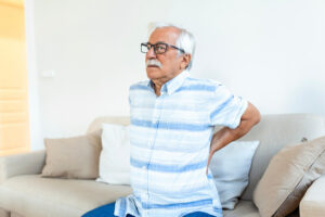An elderly man is experiencing lower back pain while sitting on his couch at home.