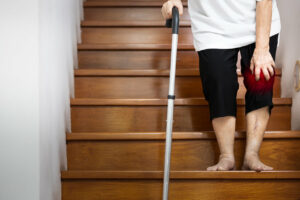 An elderly woman's hand rests on her painful knee while using a walking stick for support as she walks due to osteoarthritis.