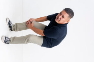 A middle aged asian man winces in pain. Bending down clutching his bum knee.
